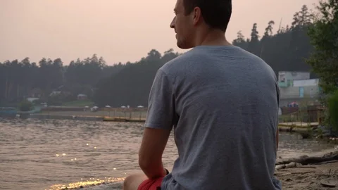 A young man is sitting against a sunset and coastal line Video stock 112952085