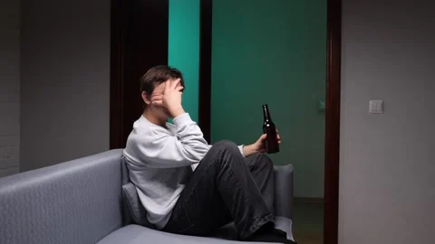 Young man sitting alone on a sofa holding an alcohol bottle, covering his face Stock Footage 346251041