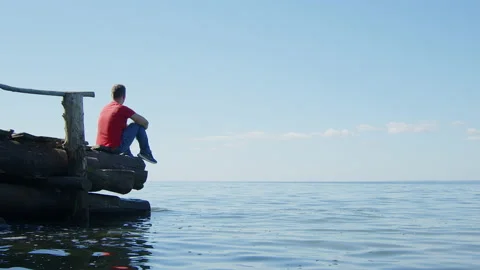 Young man sitting alone on wooden pier by sea and enjoying peace and tranquility Stock Footage 213530274