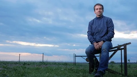 A young man is sitting and looking at the camera at sunset. A field of green Stock Footage 76918798
