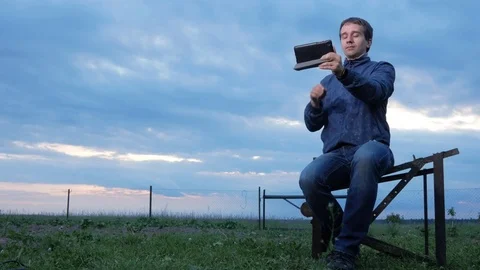 A young man is sitting and talking on the tablet at sunset. A field of green Stock Footage 76919273