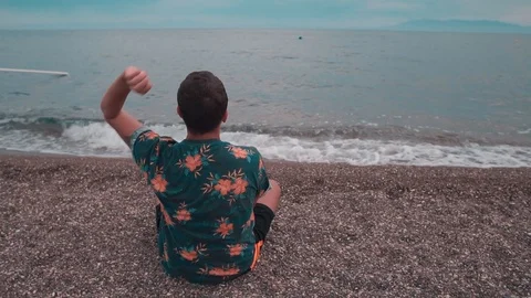 Young man sitting on the beach and  throwing pebbles into the sea.  스톡 동영상 109292550