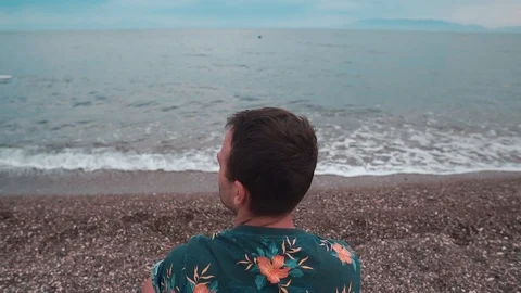 Young man sitting on the beach and  throwing pebbles into the sea. 스톡 동영상 109292555