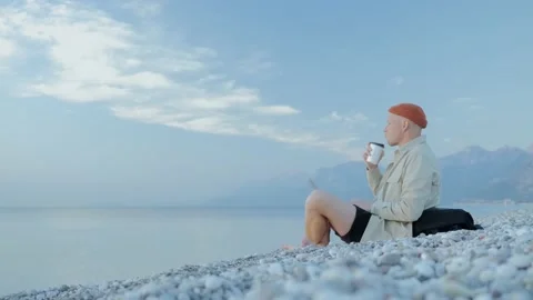 A young man is sitting on the beach and drinking a hot coffee or tea Video stock 221583725