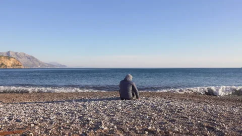 Young man sitting on the beach close to the blue sea and throwing pebbles Stock Footage 104942606