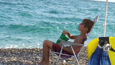 Young man sitting on the beach eating chips Vidéo 80447336