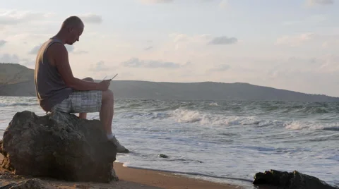A Young Man Sitting on the Beach With a Laptop on the Rocks. the Waves Beat Stock Footage 66782852
