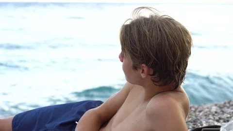 Young man sitting on beach watching waves and eating sandwich, Vidéo 79902959