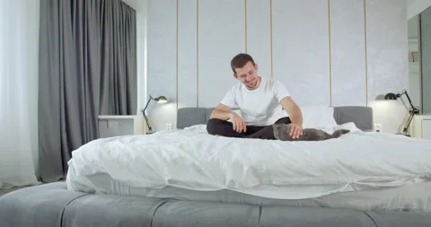 Young man is sitting on the bed and petting his cat, modern apartment, 4k Stock Footage 135376230