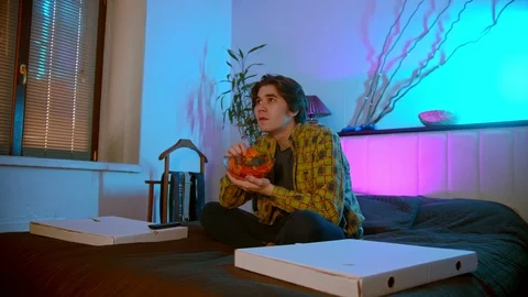 Young man sitting on the bed watching scary movie and eating chips Stock Footage 129045718