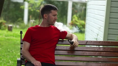 Young  man sitting on a bench and smoking electronic cigarette in the backyard Stock Footage 113183936