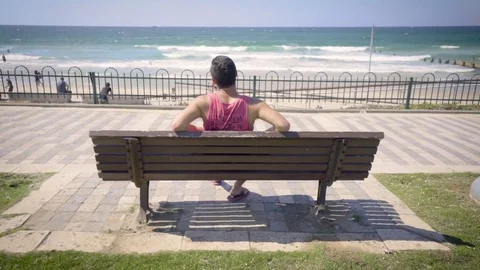 Young man sitting on bench overlooking beach Video stock 81207831