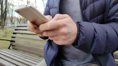 Young man is sitting on a bench in park and typing a text message in his phone Stock Footage 188973146