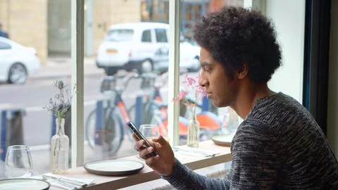 Young Man Sitting In Cafe Window Seat Checking Messages Or Social Media Using 写真素材