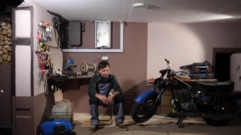 Young man sitting on a chair, pausing from his work on a vintage motorcycle in Stock Footage 342773406