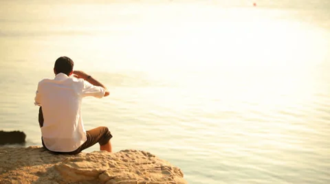 A young man sitting on a cliff looking out into the ocean during magic hour Stock Footage 33847673