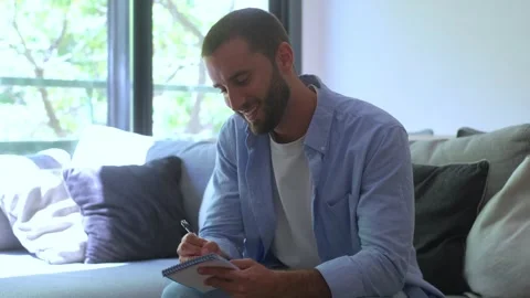 Young man sitting on a couch and writing in a notebook, smiling as he works on Stock Footage 344782288