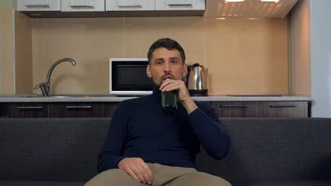A young man sitting on the couch, drinking beer and watches entertainment on TV Stock Footage 121840445