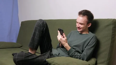Young man sitting on a couch engaged in conversation on a smartphone Stock Footage 301522063