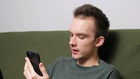Young man sitting on a couch engaged in conversation on a smartphone Stock Footage 301522066