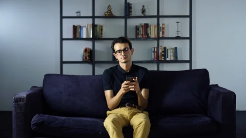 Young Man Sitting on the Couch at Home Texting on a Phone, Using New Technology Stock Footage 93256332