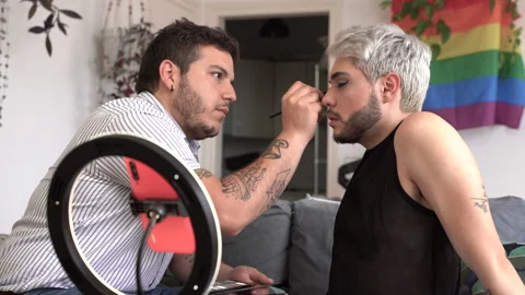 Young man sitting in the couch at home applying eyeshadow to his boyfriend. T Stock Footage