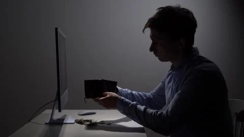 Young man sitting at a desk in a dark room, holding an empty wallet, feeling Stock Footage 342671530