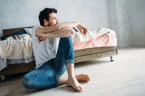 Young man sitting down on the floor near the bed and looking outside Stock Photos