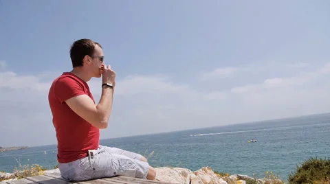 Young man sitting on the edge of the cliff and eats sandwich Stock Footage 64960646