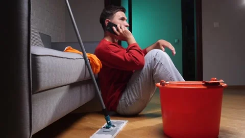 Young man sitting on the floor, calling using a mobile phone next to a mop and Stock Footage 342764942