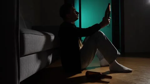 Young man sitting on the floor in a dark room with a bottle of beer, struggling Stock Footage 342771996