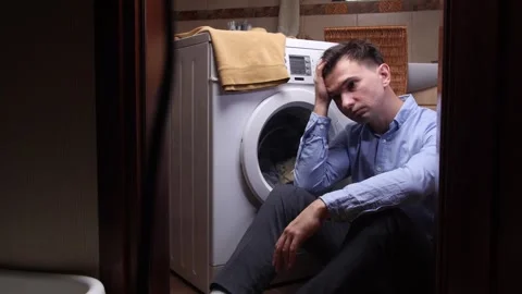 Young man sitting on the floor in front of a domestic washing machine, feeling Stock Footage 324768365