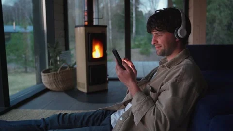 Young man is sitting on the floor by the sofa with headphones and a phone in his Stock-Footage 245988340
