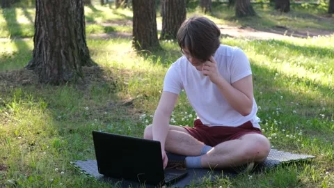 Young man is sitting on the grass in park with his laptop and talking on the Stock Footage 152222943