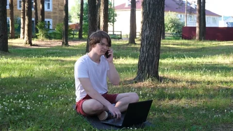 Young man is sitting on the grass in park with his laptop and talking on the Stock Footage 152223041