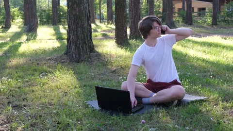 Young man is sitting on the grass in park with his laptop and talking on the Stock Footage 153532091