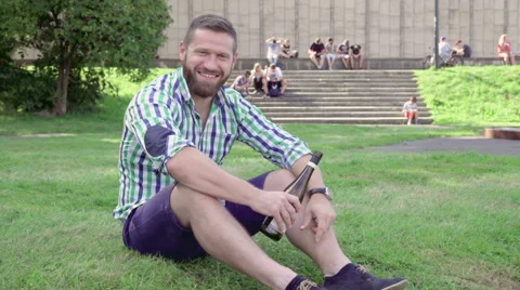 Young man sitting on grass, tastes wine from bottle and looks to camera. 库存影片 67239152