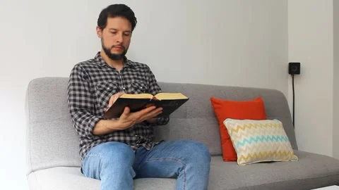 Young man sitting on a grey couch reading the holy bible Vídeo Stock 340583385