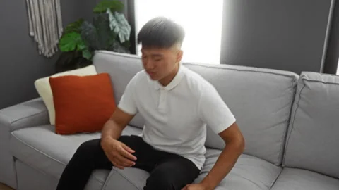 Young man sitting on a grey sofa in a modern living room, looking upset and.. Stock Footage 301095577