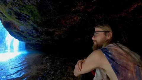 Young man sitting in grotto under waterfall Stock Footage 115093191
