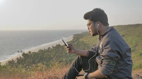 Young man sitting on a hill top browsing and listening to music Stock Footage 89952286