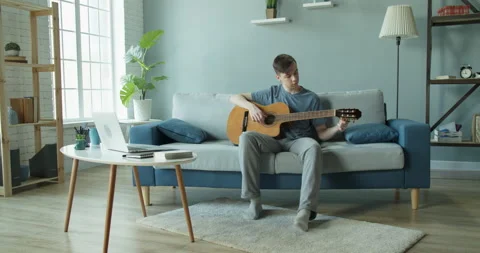 A young man is sitting at home on the sofa and tuning a guitar. Stock Footage 136296468
