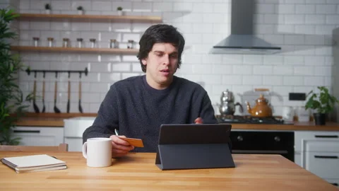 Young man sitting in kitchen at home entering credit card number on tablet .. Stock Footage 261229337
