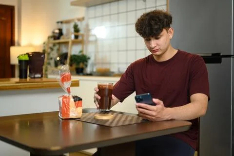 Young man sitting at kitchen table drinking chocolate protein shake and using Stock Photos
