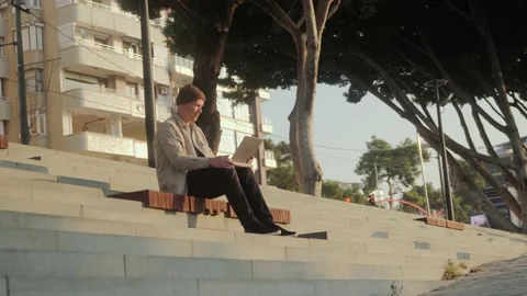 A young man is sitting with a laptop on the steps early in the morning at dawn Stock Footage 220851837