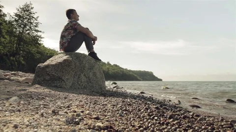 A young man is sitting on a large rock by the sea. Video stock 78700879