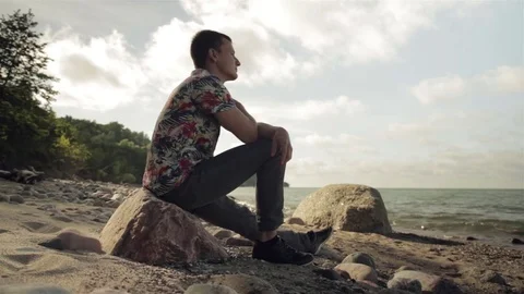 A young man is sitting on a large rock by the sea. Stock Footage 78705748