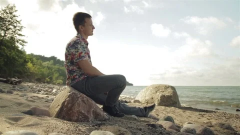 A young man is sitting on a large rock by the sea. Vídeos de archivo 78711776