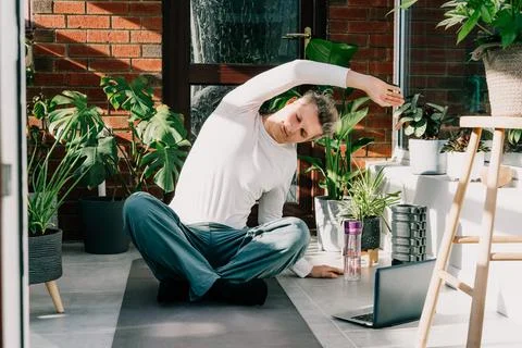 Young man sitting on mat, doing stretching yoga position in cozy modern loft Stock-Fotos