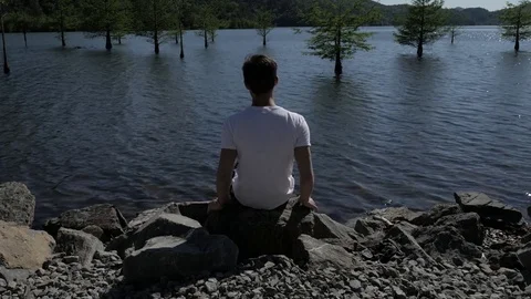 A young man sitting on rocks looking out at a beautiful river Stock Footage 75388505